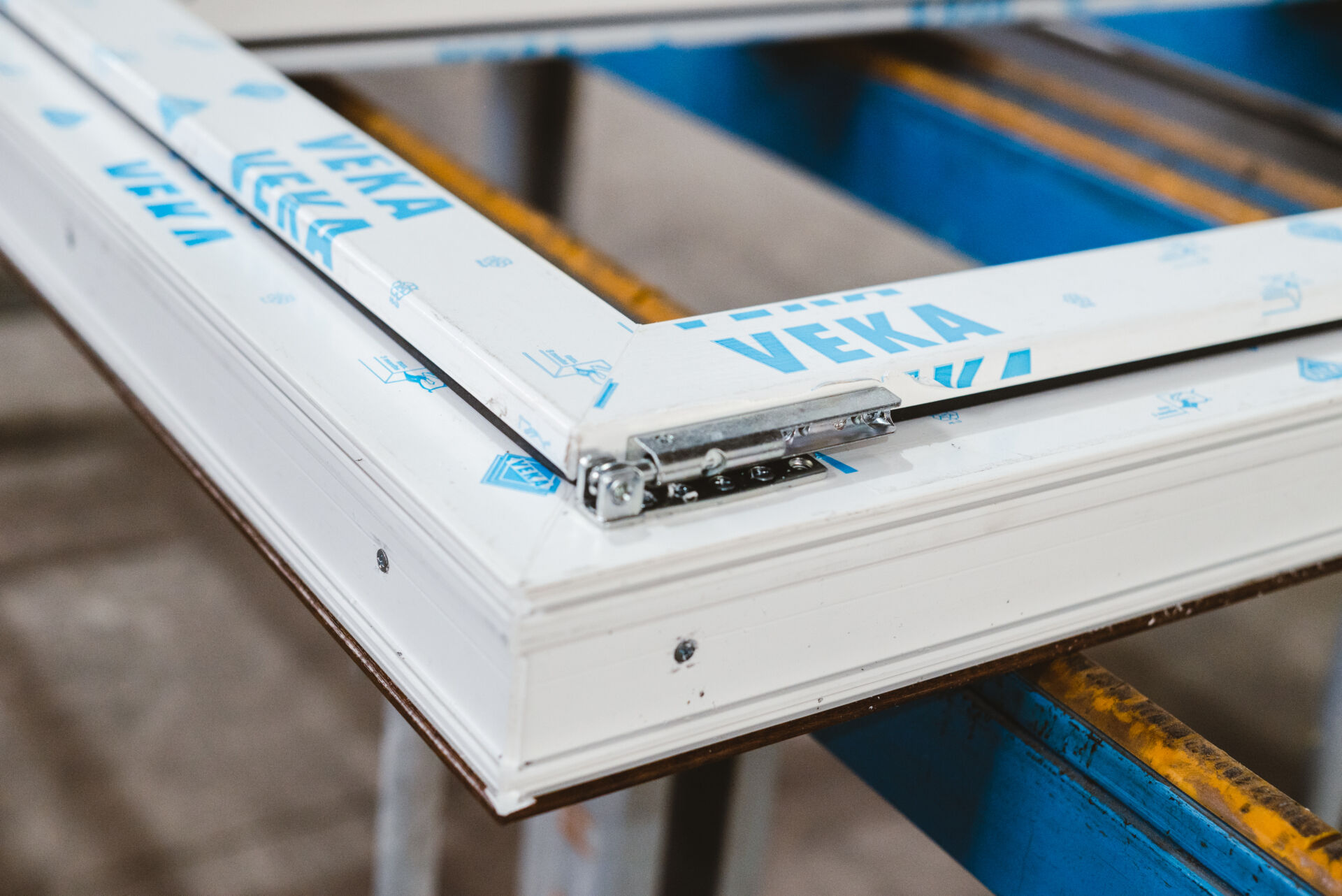 Angolo di telaio bianco in PVC con pellicola protettiva VEKA azzurra e cerniera metallica su banco blu e giallo